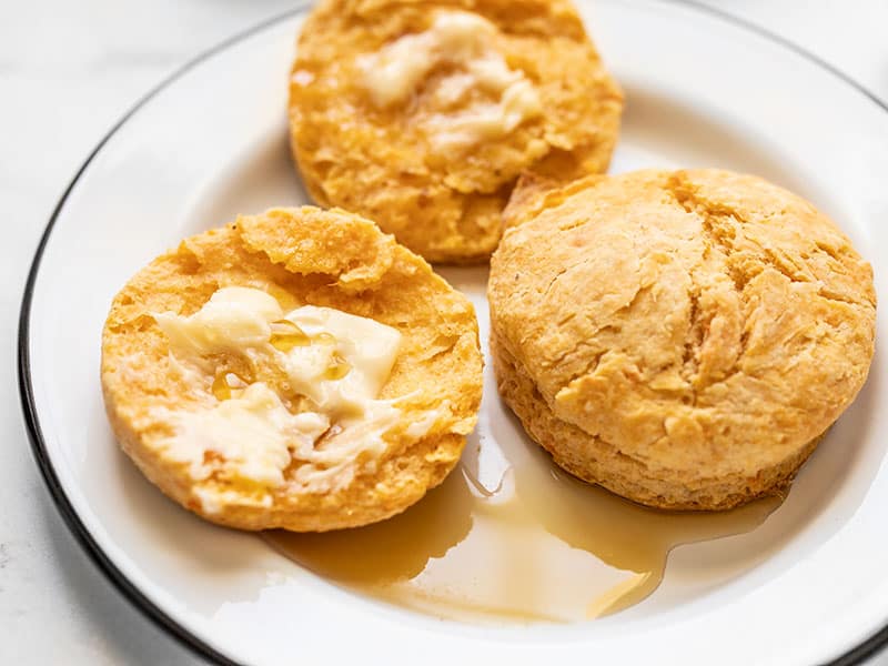 Close up of two sweet potato biscuits on a plate, one open, smeared with butter, and drizzled with maple syrup